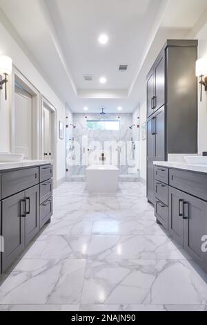 Ampia vista angolare del bagno principale in una casa moderna con piastrelle di marmo bianco, vasca da giardino, cabina doccia e armadietti di legno scuro Foto Stock