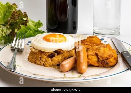 Vista laterale del riso fritto americano in stile tailandese con uova fritte, salsicce, uva passa di pollo fritta e verdure verdi in piatto di ceramica bianca con metallo Foto Stock