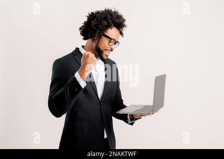 Eccitato fortunato afroamericano uomo d'affari in vestito nero e occhiali guardando lo schermo del computer portatile e celebrando la vittoria isolato su bianco, l'uomo che celebra il successo, ha fatto un buon affare Foto Stock