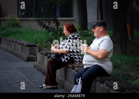 Immagine di due anziani, anziani e donne, seduti, rilassandosi e mangiando cibo e caffè da asporto per le strade di Caransebes, Romania. Foto Stock
