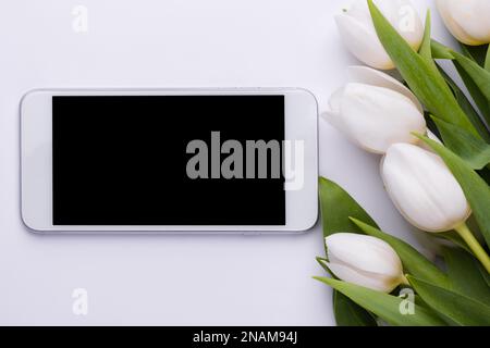 Telefono con schermo vuoto e un bouquet di tulipani bianchi. Smartphone mock up. Biglietto d'auguri di primavera online digitale. Foto Stock