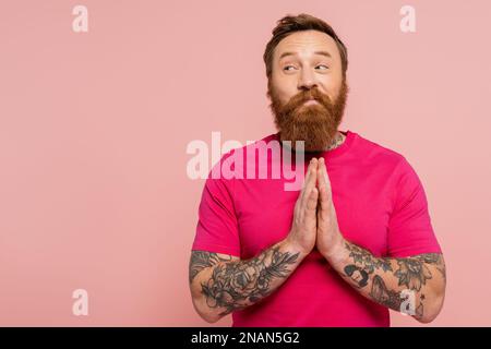 uomo sorridente e ingannevole in t-shirt magenta mostrando prego gesto e guardando lontano isolato su rosa, immagine stock Foto Stock