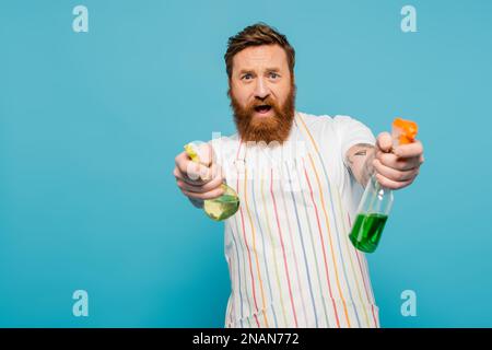 uomo eccitato in grembiule divertirsi mentre spruzzare detergenti a macchina fotografica isolato su blu, immagine stock Foto Stock