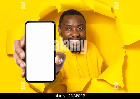 Raccomandazione app. Uomo nero felice che mostra il grande smartphone con schermo vuoto attraverso lacerato giallo foro carta, mockup Foto Stock