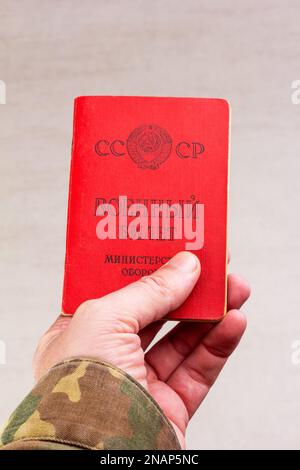 Una carta d'identità militare sovietica è tenuta in mano a un uomo in uniforme militare, in primo piano su uno sfondo grigio Foto Stock