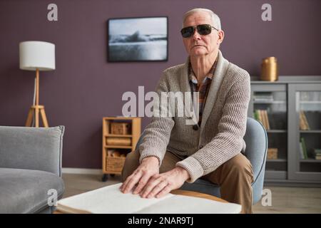 Un uomo anziano cieco negli occhiali che legge un libro in Braille al tavolo nel soggiorno Foto Stock