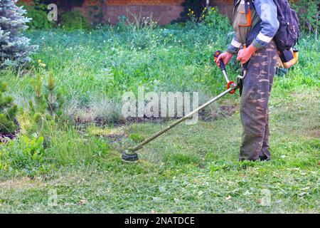 Un giardiniere taglia un prato verde in eccesso vicino alla parete della casa con un trimmer a benzina. Spazio di copia. Foto Stock