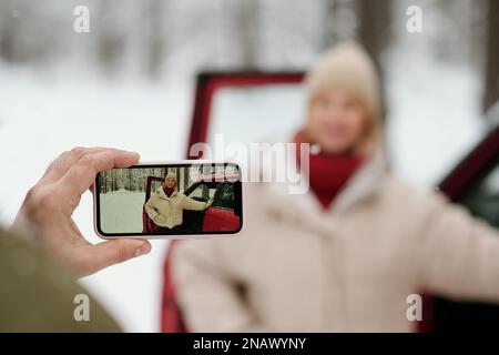 Attenzione selettiva alla mano di un uomo maturo che tiene lo smartphone mentre fotografa sua moglie in inverno in piedi dalla porta aperta dell'auto Foto Stock