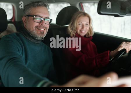 Felice uomo maturo in occhiali e caldo maglione verde che mostra la mappa locale in smartphone a sua moglie tenendo il volante durante la guida Foto Stock