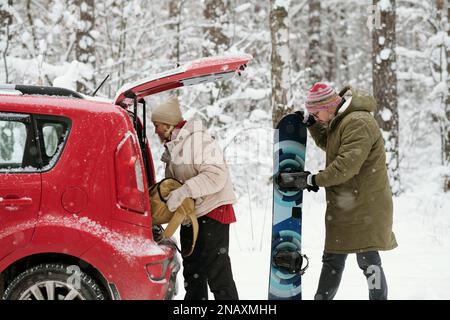 Vista laterale di una coppia matura attiva in inverno che porta il bagaglio fuori dal bagagliaio della vettura di hatchback mentre un uomo anziano guarda lo snowboard Foto Stock