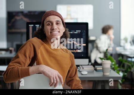 Giovane manager sorridente del supporto IT seduto sul posto di lavoro davanti alla telecamera, contro il monitor del computer con i dati grafici sullo schermo Foto Stock