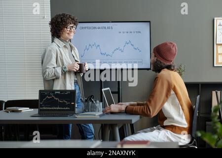 Felice giovane economista femminile con una tazza di caffè che parla con un collega maschile in casualwear seduto sul posto di lavoro di fronte a lei Foto Stock