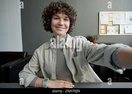 Donna d'affari sorridente brunette con capelli ricci corti e scuri che guardano la macchina fotografica mentre tiene il gadget mobile davanti a se stessa Foto Stock