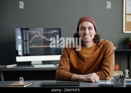 Giovane uomo sorridente broker in casualwear guardando la fotocamera mentre si siede sul posto di lavoro contro lo schermo del computer con dati grafici finanziari Foto Stock