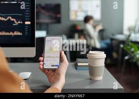Primo piano di un giovane economista o commerciante maschile che tiene uno smartphone con dati grafici sullo schermo mentre è seduto sul posto di lavoro in ufficio Foto Stock