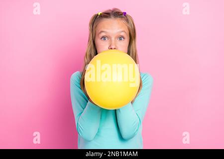 Primo piano foto di giovane carino piuttosto scolaro preteen ragazza indossare cyan pullover soffio giallo aria palloncino preparazione evento isolato sul colore rosa Foto Stock