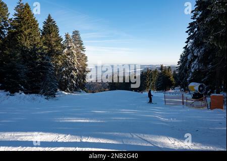 Pista da sci sul Wasserkuppe con sciatori, cannone da neve sul lato e con cielo blu e alberi Foto Stock