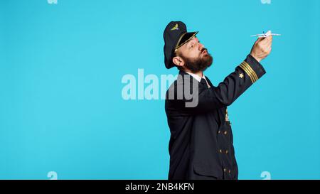 Uomo caucasico vestito come pilota che gioca con un giocattolo per aeroplano, mostrando un piccolo velivolo artificiale in miniatura sulla macchina fotografica. Giovane adulto che lavora come capitano volante con finto mini aereo su sfondo blu. Foto Stock