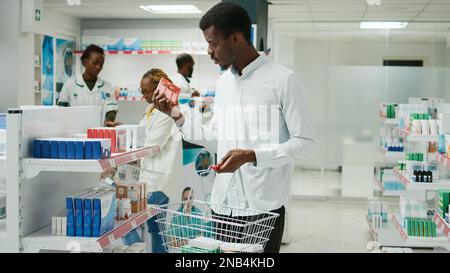 Cliente afroamericano mettere farmaci nel cestino, alla ricerca di vitamine e bottiglie di pillole. Giovane cliente maschio che acquista trattamento di prescrizione e supplementi, farmacia negozio. Foto Stock