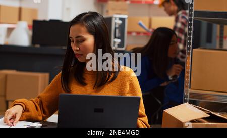 Dipendente asiatico che lavora sulla logistica delle scorte con il laptop, pianificando la spedizione degli ordini per lo sviluppo aziendale in magazzino. Donna che gestisce la supply chain alla scrivania, al dettaglio. Scatto manuale. Foto Stock