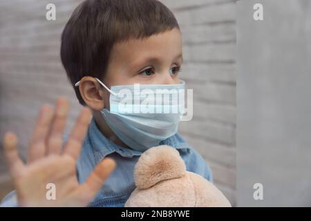 Triste ragazzino in maschera protettiva che guarda fuori dalla finestra interna, vista dall'esterno. Rimanere a casa durante la pandemia di coronavirus Foto Stock