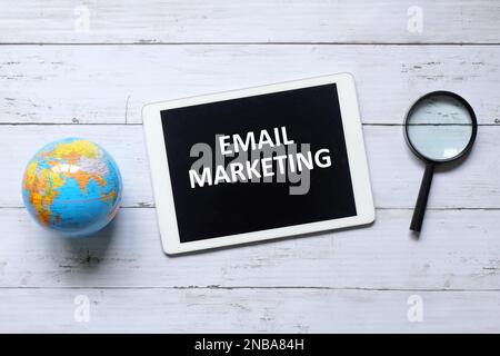 Vista dall'alto della mappa del globo del mondo, lente di ingrandimento e tablet scritto con "E-MAIL MARKETING" su sfondo bianco di legno. Foto Stock