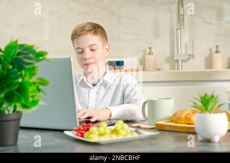 giovane ragazzo, adolescente gioca sul notebook e sul computer portatile durante la colazione. Dipendenza dai gadget. Foto Stock