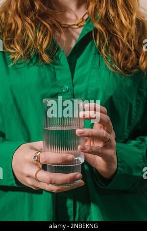 Primo piano di una donna irriconoscibile in una camicia verde brillante con vetro fluted trendy e acqua pulita. Foto Stock