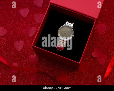 Confezione regalo con orologio argento è su sfondo rosso cuore carta, vista dall'alto. Biglietto d'auguri, presente. Concetto di vacanza di San Valentino. Appartamento con un elegan Foto Stock