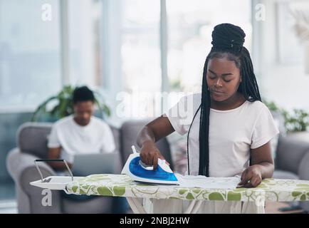 Appena pressato e senza grinze. una giovane donna che stira abiti con il marito sul divano sullo sfondo. Foto Stock
