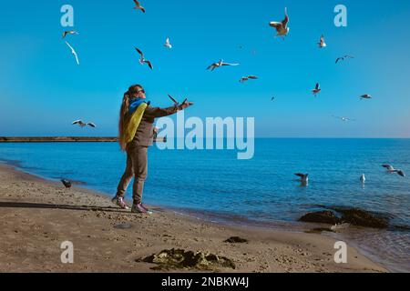 Bella donna matura con la bandiera Ucraina sulle sue spalle si nutre gabbiani con lo sfondo del cielo blu e del mare in Odessa Ucraina Foto Stock