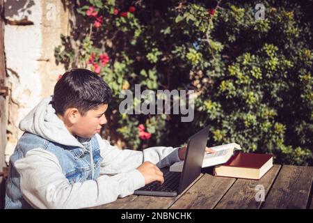 il giovane uomo latino legge il libro davanti al notebook, all'aperto Foto Stock