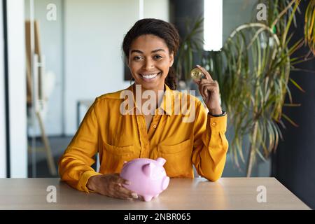 Felice manager femmina che mette la moneta bitcoin in banca piggy all'interno dell'ufficio, donna sorridente alla macchina fotografica, seduto alla scrivania Foto Stock