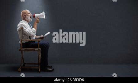 Regista professionista e sicuro seduto sulla sedia del regista e gridando con un megafone, concetto di industria cinematografica Foto Stock