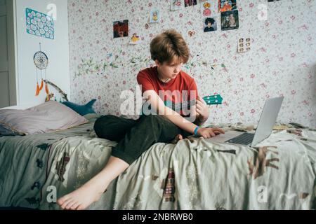 Ragazza adolescente seduta a letto imparare a giocare ukulele utilizzando lezioni online sul computer portatile. Foto Stock