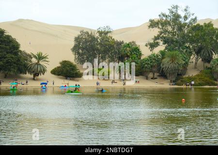 La gente salite in barca al lago del villaggio di Huacachina con palme e colline di sabbia. Messa a fuoco selettiva. Foto Stock