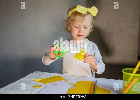 Un bambino piccolo sorride mentre taglia i dettagli per una carta del giorno della mamma e il 8 marzo. La ragazza fa un regalo a sua madre. Foto Stock