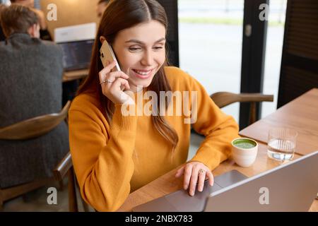Primo piano di una bella donna d'affari di brunette caucasica sorridente che parla sul suo smartphone con un amico mentre lavora su un computer portatile in remoto. Foto Stock