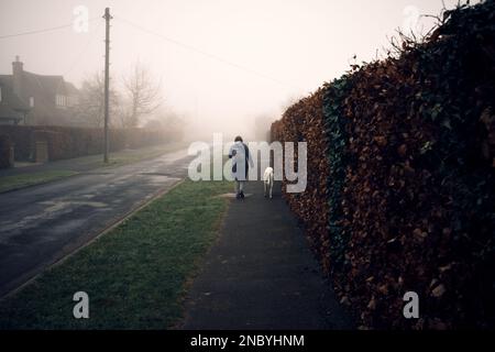 Da dietro, una donna cammina un cane levriero in una giornata misteriosa e oscura in un villaggio del North Yorkshire. Foto Stock