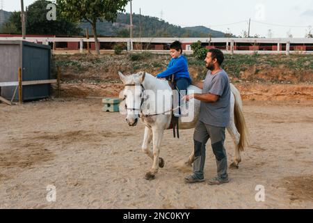 Bambino con disabilità che fa un giro a cavallo mentre fa una sessione di terapia equina. Foto Stock