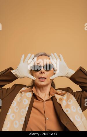 sorpresa donna anziana in occhiali da sole e vestito posa isolato su beige Foto Stock