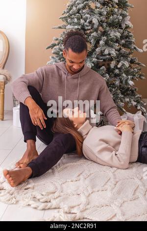 Una ragazza felice, si trova in grembo di un ragazzo alla moda vicino all'albero di Natale nella stanza Foto Stock