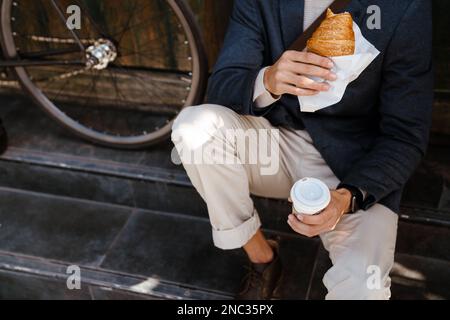 Primo piano di un uomo che tiene una tazza di caffè e croissant seduti vicino alla bicicletta all'aperto sulla strada cittadina Foto Stock