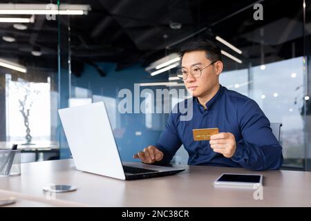 Un giovane banchiere asiatico lavora in ufficio in un computer portatile, ha un problema di credito nelle sue mani. Consiglia, parla, offre un account tramite videochiamata ai clienti. Foto Stock