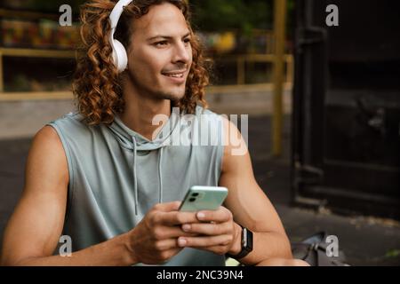 Ritratto corto di giovane uomo sportivo sorridente a capelli lunghi in cuffie tenendo il telefono e guardando da parte Foto Stock