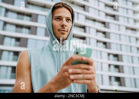 Giovane atletico uomo bello dai capelli lunghi in cuffie e cappuccio tenendo il telefono e guardando da parte, mentre si trova vicino a un edificio moderno all'aperto Foto Stock