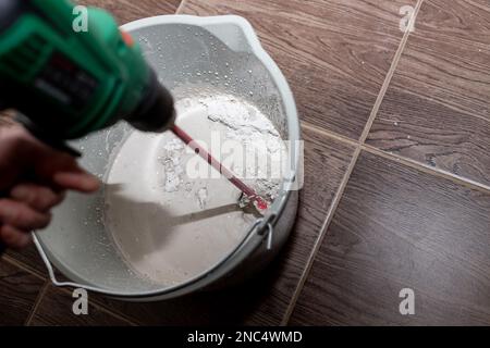 lavoratore che mescola intonaco in un secchio con trapano elettrico. Un primo piano di tessitura dell'intonaco. L'uomo mescola la Malta con un perforatore con un ugello speciale Foto Stock