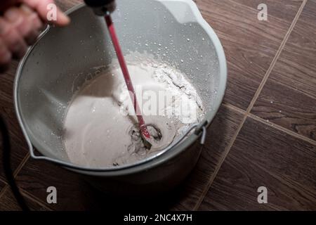 lavoratore che mescola intonaco in un secchio con trapano elettrico. Un primo piano di tessitura dell'intonaco. L'uomo mescola la Malta con un perforatore con un ugello speciale Foto Stock