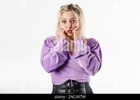 Ritratto di donna bionda che indossa blusa in piedi isolato su mani di sfondo bianco in pugni, toccando mento, guarda la bocca WOW alla macchina fotografica ed è du Foto Stock