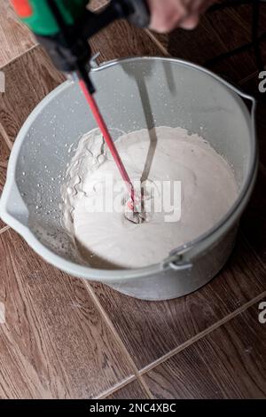 lavoratore che mescola intonaco in un secchio con trapano elettrico. Un primo piano di tessitura dell'intonaco. L'uomo mescola la Malta con un perforatore con un ugello speciale Foto Stock